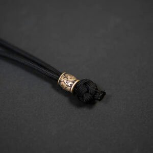 Žalvarinis rankų darbo karoliukas „Vytis“ su paracord virve, cilindro formos, pritvirtintas prie juodos virvės mazgo, fotografuotas ant tamsaus fono.