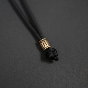 Žalvarinis rankų darbo karoliukas „Gedimino Stulpai“ su paracord virve, cilindro formos, pritvirtintas prie juodos virvės mazgo, fotografuotas ant tamsaus fono.