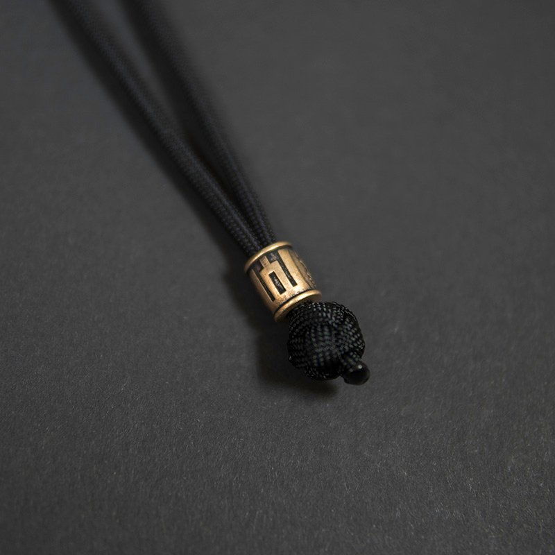 Žalvarinis rankų darbo karoliukas „Gedimino Stulpai“ su paracord virve, cilindro formos, pritvirtintas prie juodos virvės mazgo, fotografuotas ant tamsaus fono.